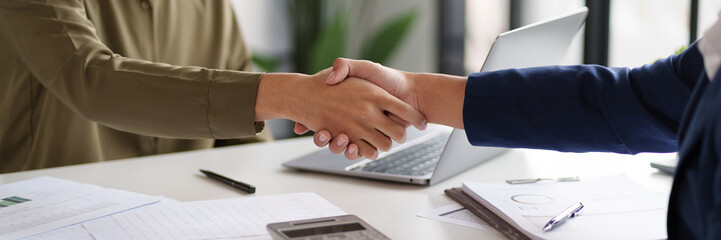 Two individuals are shaking hands across a desk, indicating a successful agreement or partnership....