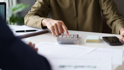 A professional setting featuring two individuals working at a table. They are engaged in reviewing documents and using a calculator. Laptops are open on the table.