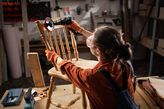 Woman using sander with a metal round bristle disk in a carpenter's workshop, sanding a wooden chair in the process of producing