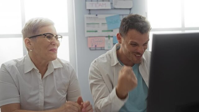 Woman and man working and laughing together in an office setting with a computer screen and posters on the wall in the background, portraying a lively and cooperative workplace environment