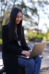 A woman is sitting on a bench with a laptop in front of her. She is wearing a black coat and blue jeans. The scene suggests a casual and relaxed atmosphere, as the woman is using her laptop outdoors