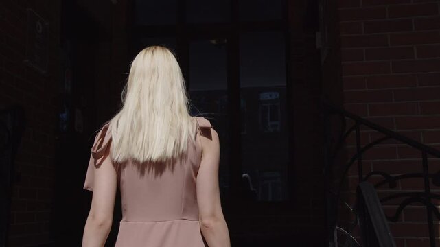 Blonde in a pink long dress and carrying a large leather bag walks up the stairs