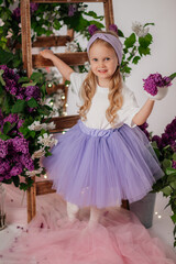 A little blonde girl, with a mischievous grin, holds a unique cup filled to the brim with vibrant lilac blossoms. 