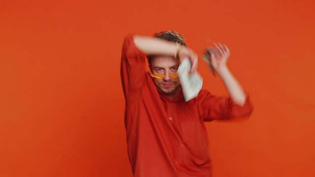 Happy rich Caucasian man holding fan of cash money dollar banknotes celebrate dancing, success business career, lottery game winner, big income, wealth. Young guy isolated on orange background indoors