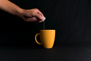 Latin man's hand. The hand is taking out a tea bag from a yellow cup on black background. Yellow mug. Corporate color. ceramic mug