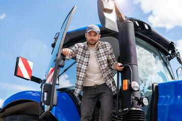 Naklejka premium Portrait Caucasian ethnicity man farm tractor driver in plaid shirt and cap on blue tractor.