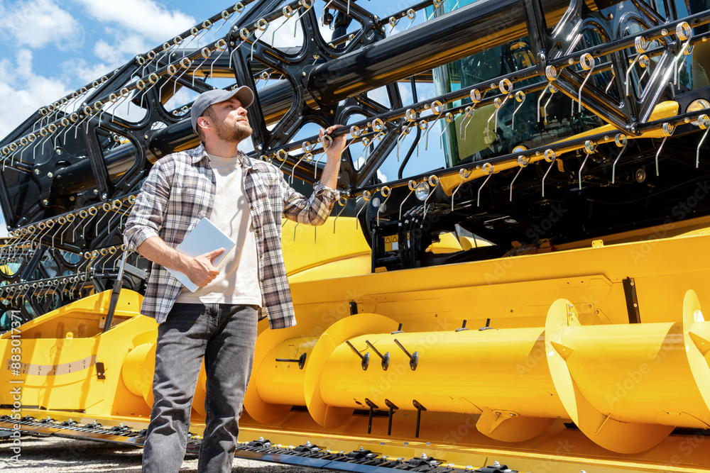 Wall mural caucasian adult man with digital tablet in hand looking at a yellow combine harvester at a farm mach