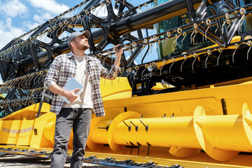 Caucasian adult man with digital tablet in hand looking at a yellow combine harvester at a farm...