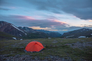 Orange tent on grassy stony pass with view to mountain range silhouette far away under clouds in vivid sunset color sky. Scenic alpine landscape with red tent in mountains under colorful cloudy sky.