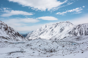 Cold snow-white landscape of snowy valley with alpine lakes against snow-covered rocky mountain range under clouds in blue sky. Glacial lakes, pure white snow and stones in high mountains in sunny day