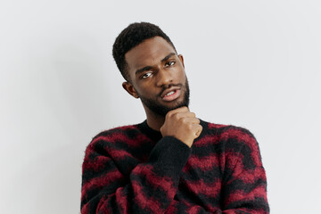 Contemplative young man in stylish red and black sweater standing against a white wall with hand on chin