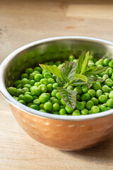 Petits pois peas in a capper serving bowl with freshly picked mint garnish. On a wooden cutting board.