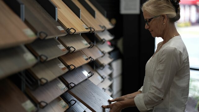 Happy mature woman looks at solid or engineered wood plank samples at a display in a floor or hardware store for a home remodeling renovation project.