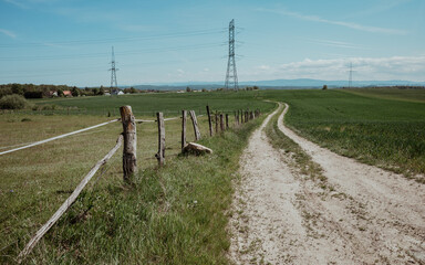 Peaceful summer afternoon, a rural landscape unfolds, featuring a wooden fence alongside a sandy road stretching to the horizon amid green pastures. In the distance, tall power poles are visible