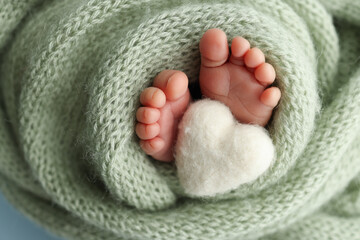 The tiny foot of a newborn baby. Soft feet of a new born in a olive green wool blanket. Close up of toes, heels and feet of a newborn. Knitted white heart in the legs of a baby. Macro photography. 