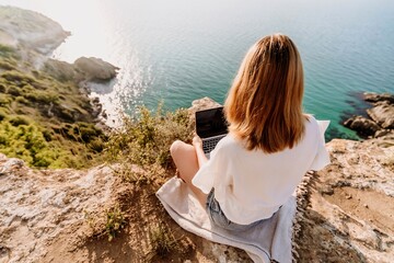 A woman is sitting on a rock overlooking the ocean with a laptop in front of her. She is enjoying the view and the peacefulness of the location.