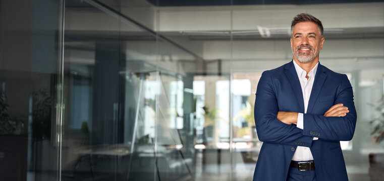Handsome smiling confident hispanic senior man with crossed arms looking aside. Indian or latin successful mature middle age leader male businessman, business investor in office. Banner, copy space.