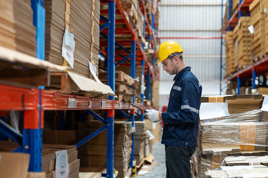 Man warehouse worker wearing uniform and helmet safety standing with clipboard for checking stock products on shelves in warehouse