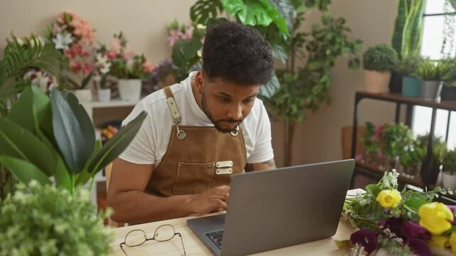 Focused black man using a laptop in a vibrant flower shop filled with various plants and floral arrangements indoors.