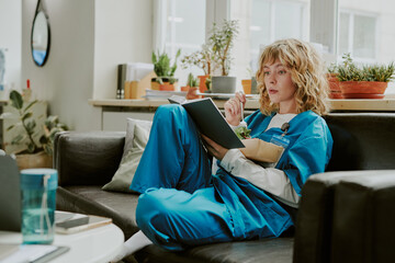 Young blond Caucasian nurse eating stabbing fresh salad while reading novel with enthusiasm
