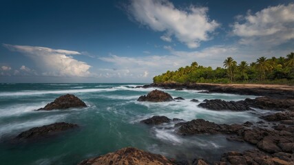 A stunning tropical coastline featuring rocky shores, gentle waves, and lush palm trees under a vibrant blue sky with clouds.
