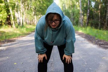 With her hands on her knees, a woman catches her breath after a long run. Sweat runs down the face of a plump woman after a cardio workout by running. A woman rests after a run.