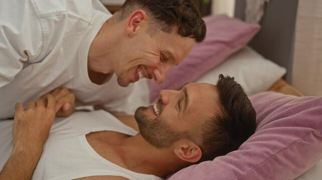 Couple laughing together in bed at home with purple pillows in the bedroom celebrating their gay love and commitment to each other