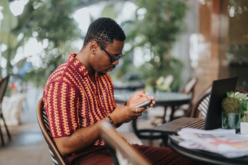 Young man concentrating on his smart phone while working on a laptop at an outdoor cafe. Modern technology, remote work, and casual work environment concept.