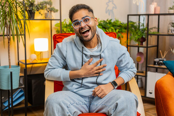 Happy excited Indian man laughing out loud after hearing ridiculous anecdote, funny silly joke, feeling carefree amused, positive people lifestyle at modern home room apartment. Guy sits on chair