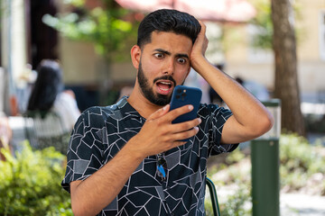 Young bearded Indian man use smartphone typing browsing, loses becoming surprised sudden lottery...