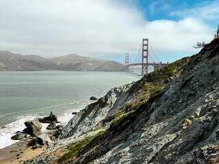 Coastal view of the Golden Gate bridge in San Francisco. American iconic landmark. Cliffs and grass frame the picture. Waves in the bay water. Light fog.