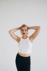 A blonde woman in a white crop top and black pants poses gracefully with arms extended upwards, set against a plain white background.