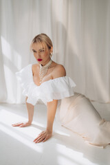 Elegant woman with blonde bob haircut and red lipstick posing on all fours in a white ruffled dress and pearl necklace, against a white background. Copy space...