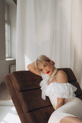  Elegant woman with blonde bob haircut and red lipstick reclining on a brown chair, wearing a white...