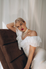  Elegant woman with blonde bob haircut and red lipstick reclining on a brown chair, wearing a white...