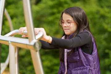 Naklejka premium A little cute smiling girl in glasses and a purple vest plays on the playground. Happy childhood concept