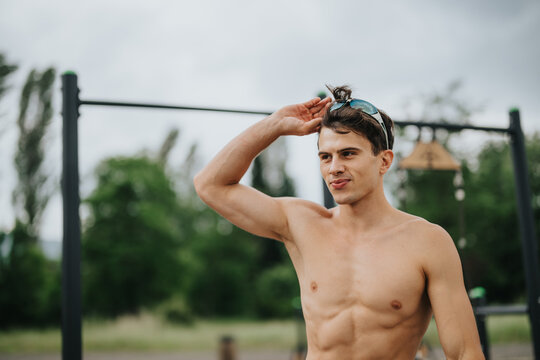 Fit young man adjusting sunglasses at an outdoor gym. Shirtless athlete standing near parallel bars on a cloudy day.