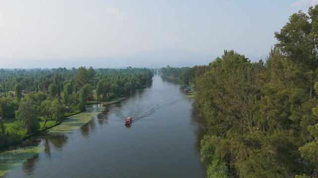 Aerial Tilt Up Video Over Xochimilco Canal in Mexico City: Red Trajinera Glides Peacefully Amid Green Trees and Surrounding Boats, Creating a Cozy Urban Oasis
