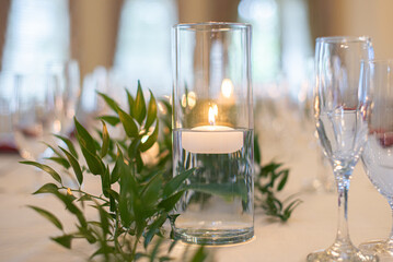 Tall glass vase with lit tea light candle floating inside on wedding reception dinner table with green plants