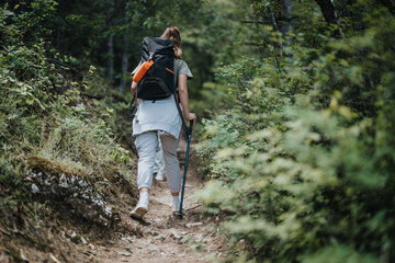 Obraz premium Back view of a woman hiking through a forest trail equipped with a backpack and trekking poles, enjoying a summer day in nature.
