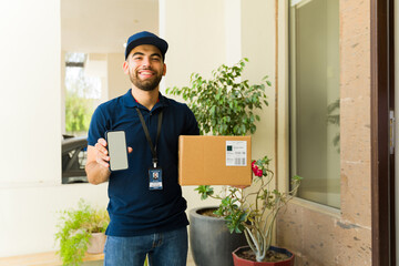 Smiling delivery man holding a package and showing a smartphone with a blank screen at a house entrance