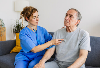 Young female doctor in blue uniform listening to disabled older patient heart sound with stethoscope during visit at home. Medical checkup and healthcare concept