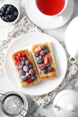 Tasty puff pastries with berries and tea on white marble table, flat lay