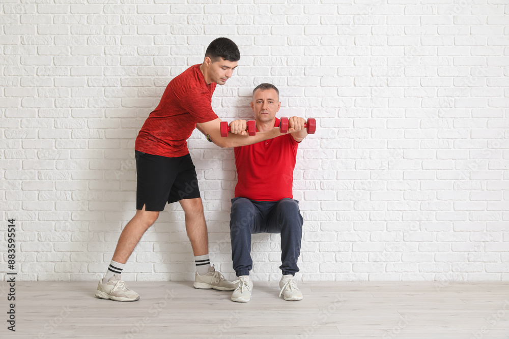 Sticker mature man training with dumbbells and rehabilitation therapist near white brick wall