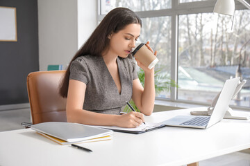 Pretty businesswoman writing on clipboard and drinking coffee at table in light office