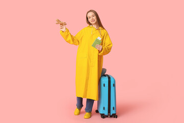 Smiling young woman in raincoat with wooden airplane and passport on pink background. Travel concept
