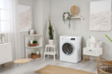 Blurred view of light laundry room with modern washing machine, houseplant and bottles of detergent