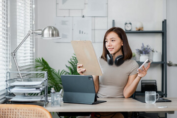 A young woman multitasking in a modern home office, using a tablet and smartphone, with headphones...