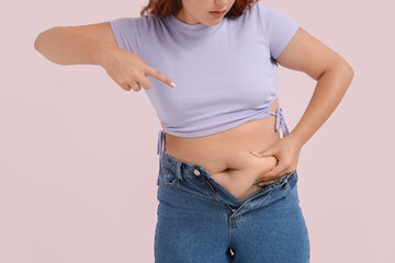 Overweight woman in tight jeans pointing at her belly on pink background. Excess weight concept