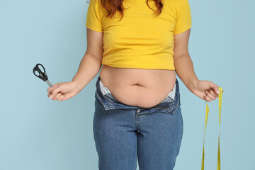 Overweight woman in tight jeans with measuring tape and scissors on blue background. Weight loss...